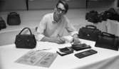 Väsk Reportage modenummer 26 oktober 1967

En ung man klädd i vitrandig skjorta och som bär glasögon sitter och designar väskor vid ett bord. Tre handväskor står på bordet och även skinnprover ligger där. Fler handväskor står på hyllor i bakgrunden.