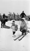 Ånnaboda 12 februari 1968

En liten flicka står i Ånnabodas skidbacke med en nalle i famnen. Hon är klädd i vit mössa med mörk tofs på huvudet, mörk halsduk, vit jacka, mörka byxor och vita stövlar. Invid henne knäböjer en kvinna klädd i vit mössa, vitmönstrad jacka, svarta byxor och svarta pjäxor. Hon har skidor på fötterna och håller ett par stavar klämda mot vänstra sidan av kroppen med vänster arm. Fler personer samt ett ställ med skidor syns i bakgrunden. En person lastar något i en pulka.