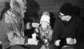 Ånnaboda 29 januari 1968

En familj fikar utomhus i Ånnaboda. Mamman till vänster matar ett litet barn med kanelbullebitar. Mamman är klädd i ljus, pälsmössa, ljusmönstrad jacka och mörka byxor. Det lilla barnet är klädd i vit mössa med tofs, ljusmönstrad jacka och ljusa byxor. I sin vänstra hand håller barnet i ett par svarta solglasögon. Pappan är klädd i mörk tröja, mörk halsduk, mörk mössa och mörka byxor. Föräldrarna har kaffekoppar i händerna. I bakgrunden syns en husvägg.