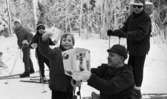 Ånnaboda, fotojobb (?), arbetarkommun 70 år 12 februari 1968

Affärsmannen Totta som äger affären Tottas sport står på knä framför en liten pojke i sjuårsåldern med en vit låda i sina händer. På lådan står det: 