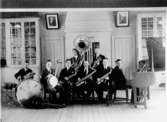 Rumsinteriör, orkester, åtta män med musikinstrument.