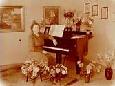 Fru Falk, pianolärarinna vid ett piano.
Rumsinteriör, högtid.