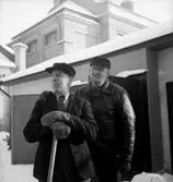 Fo-staben, två män.
Byggnader i bakgrunden.