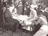 Diakonissan Valborg Kihlberg leder en kurs i sjukvård. Kursdeltagarna, unga kvinnor och män, övar sig. Några av dem har fått bandage.