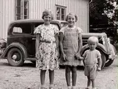 Tre barn fotograferade utomhus. I bakgrunden skymtar en bil, (Nils Andersson, 