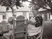 En kvinna (oidentifierad) med hushållsarbete (spritar ärtor?) utomhus i en trädgård. Vid grinden står en pojke, troligen Rolf Mattsson. I bakgrunden bostadshus och ekonomibyggnad. Fastarpsvägen, Tvååker.
