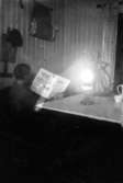 Rumsinteriör, en pojke med Nerikestidningen.
Tidningsläsning i fotogenlampans ljus.
