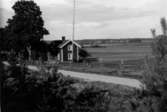 Gård i skogsbygden- mellerta gården Ö. om vägen via grindstugan. Utsikt över sjön mot Himmer. Till vänster om vägen grusbacke en del av åsen, som går förbi Lännäs kyrka. Byggnaden: mangård och ladugård. I augusti 1932