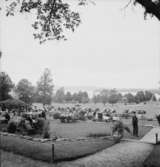 Festligheter vid Siggebohyttan den 1 augusti 1937. Folk sitter och fikar vid uteserveringen intill gågatan upp till Siggebohyttans bergsmansgård. Längre bort står bilar parkerade. I bakgrunden syns sjön Usken.