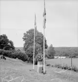 Festligheter vid Siggebohyttan den 1 augusti 1937. På grässlänten intill Siggebohyttans bergsmansgård står en talare i en talarstol och håller ett anförande. På bägge sidor om talaren står varsin flaggstång med en svensk flagga. Likaså talarstolen är iklädd en svensk flagga. Till vänster på grässlänten syns några åskådare.