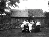 En grupp personer framför en torpstuga. En man och en kvinna sitter.  Intill paret står kvinnor och barn.