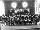 Konfirmation i Varbergs kyrka. Konfirmanderna sitter kring altarringen i kyrkans kor. Flickorna har ryggen vänd mot kameran. De har stora rosetter i håret.