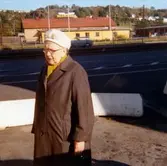 En glasögonprydd dam, klädd i brun kappa och vit hatt som står på Göteborgsvägen, år 1972. I bakgrunden syns spårvagnsspår, järnvägsspår och Ågatan.