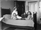 Sjukvård. Sjuksyster med patienter. Sjuksystern sitter på sängen tillsammans med två flickor. En kvinna sitter i en fåtölj.