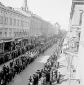 Förstamajdemonstration i Örebro den 1 maj 1937. Demonstrationståget går i riktning söderut på Drottninggatan. Det mycket långa förstamajtåget består av människor av olika slag. Några bär på röda fanor, plakat och banderoller. Åskådare kantar Drottninggatans båda trottoarer för att titta på demonstrationståget. Nikolaikyrkans torn kan skymtas ovanför hustaken till vänster i bilden. Närmast till höger står en kvinna på en balkong och tittar på evenemanget.