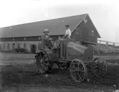 En man och en pojke på en traktor.
Ekonomibyggnader i bakgrunden.