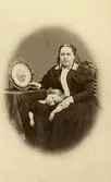 En kvinna och en liten flicka.
Doktorinnan HILMA Wilhelmina Ingelson (1831-1901), född Uddén, med dottern Helfrid.
Hilma Wilhelmina Uddén, född 1831-12-09 i Stenkvista (D), gift 1862 (?) i Kumla med WILHELM Anders Ingelson, född 1822, provinsialläkare i Tanum, Bohuslän, död 1868 i Hoghem, Tanum, O.
Dottern: Helfrid Ingelson, född 1866-05-08 i Hoghem, Tanum, gifte sig med läkare Fredrik Bissmark, som avancerade till medicinalråd.
Eftersom Hilma blev änka 1868 kan hon ha flyttat till sin bror Axel Uddén i Strängnäs. Han gifte sig 1868 när han hade fått tjänst som läroverksadjunkt där.
Fotografiet måste vara tagen runt 1868-1869, med tanke på att hon är änka på bilden och förmodligen sitter med ett porträtt av sin döde make, och flickan ser ut att vara i 2-3 årsåldern.