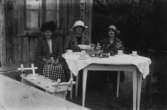 Kaffebjudning utomhus, tre kvinnor vid bordet.
Bostadshus i bakgrunden.
Augusta Olsson, Kally Vedenbrand, Thea Elfrida Olsson.