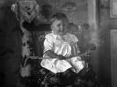 Barnporträtt. Ett barn fotograferat inomhus sittande i en stol. Framför barnet är ett rep spänt för att hindra att det faller ur.