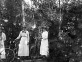 Bild av några kvinnor fotograferade på cykelutflykt i skogen. De bär långaklänningar och hatt; en av dem har en stor halsrosett.