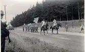 Kulturhistoriska festtåget och festspelen i Falköping den 5 augusti 1934. I täten av tåget rider två fanfarblåsande trumpetare, klädda som pager från Gustaf II Adolfs tid. Därefter följer svenska flaggan med en scoutgrupp som fanborg. Scouterna bär devisen 