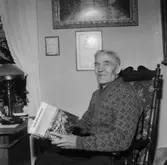 En 80-årig man i Hakarp, Huskvarna sitter med en bok i handen ( Hylte bruks aktiebolag). På väggen bakom honom hänger ett diplom med namnet August Svensson.