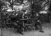 Tre män som sitter utomhus vid ett bord och dricker öl och sockerdricka, ”vy 1920” på asken
	Metallutfällning.