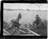 John Bauers Lapplandsresa sommaren 1904. Två samer bredvid sin båt på stranden vid en fjällsjö. De tillreder fisken de fångat.
