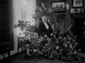 Man poserar bland blomsteruppsättningar, (födelsedag?) i rumsinteriör ”Åkare Kristersson”.