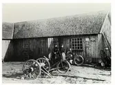 Smedja. Gesällerna Gustav och John utanför Anders Anderssons smedja i Veddige 1925. Vid huset står en motorcykel och i förgrunden ett par vagnar.