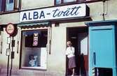 En kvinna står i dörren till tvätteriet ALBA-tvätt med adress Kvarnbygatan 43 vid Gamla Torget i Mölndal, 1960-tal. Till höger ses även en blå telefonkiosk.

För mer information om bilden se under tilläggsinformation.