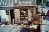Två män fotograferade utanför en blomsteraffär i huset Roten M3 vid Götaforsliden i Mölndal, 1960-1970-tal.

För mer information om bilden se under tilläggsinformation.