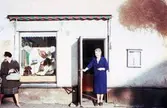 Två kvinnor står utanför Elfrida Isakssons modeaffär med adress Kvarnbygatan 45 i Mölndal, 1960-1970-tal.

För mer information om bilden se under tilläggsinformation.