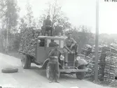 Vedtransport i trakten av Solstadström.  Stående på marken Gottfrid Andersson, på taket Erik Svensson, och på motorhuven Hjalmar Karlsson. De båda sistnämda var chaufförer. Reservhjulet är tillfälligt placerat på vägen, ringa trafik. och förvarades under körning alltid uppe på lasten.