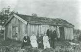 Familj fotograferad framför mycket enkel liten stuga.