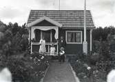En stuga, man, kvinna och barn i Kalmar södra koloniområde, fotograferat omkring 1930. Kalmar södra koloniförening grundades 1917 och har idag 105 kolonilotter. Området ligger strax söder om länssjukhuset i Kalmar med huvudingång från Stensbergsvägen.