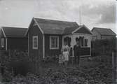 En stuga, man, kvinna och barn i Kalmar södra koloniområde, fotograferat omkring 1930. Kalmar södra koloniförening grundades 1917 och har idag 105 kolonilotter. Området ligger strax söder om länssjukhuset i Kalmar med huvudingång från Stensbergsvägen.