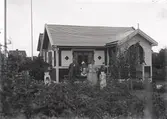 En stuga, man, kvinna och barn i Kalmar södra koloniområde, fotograferat omkring 1930. Kalmar södra koloniförening grundades 1917 och har idag 105 kolonilotter. Området ligger strax söder om länssjukhuset i Kalmar med huvudingång från Stensbergsvägen.