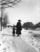 Här ses Ingeborg Malmberg, född Olsson, tillsammans med Ingrid Brundin, ca 1915-1916. I bakgrunden syns Dalapalatset och Svängbron. Snön från Gävles gator och gårdar kördes med häst och släde och stjälptes ut i Gavleån, så man kunde gå över ån på snövallarna vissa år.