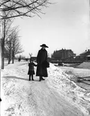 Här ses Ingeborg Malmberg, född Olsson, tillsammans med Ingrid Brundin, ca 1915-1916. I bakgrunden syns Dalapalatset och Svängbron. Snön från Gävles gator och gårdar kördes med häst och släde och stjälptes ut i Gavleån, så man kunde gå över ån på snövallarna vissa år.