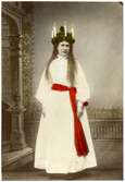 Ateljéporträtt, kolorerat, föreställande Kröök Karin Olsdotter från Leksand i Dalarna som Lucia. Hon bär långt, vitt, linne, rött band i midjan och en klädd krona med ljus. Skansens Lucia omkring 1898.