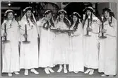 Luciafest på Sala lasarett 1921 med skämtsamt inslag. Lucia i mitten med bricka i hand omgiven av sina tärnor. Deltagarna i tåget är sannolikt personal på lasarettet som ska uppvakta patienterna.