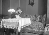 Sigrid Paues sittande i en ottoman. Läsning vid bordet. Tidsomfånget är 1900 - 1940