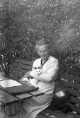 Sigrid Paues med en katt i famnen, sittande i en trädsgårdssoffa i berså. Tidsomfånget är 1900 - 1940