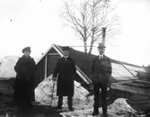 En kvinna, två män framför växthus.
