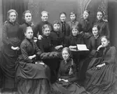 Klass 8, Högre Flickskolan. Våren 1887. Klassföreståndarinna Fröken Hanna Rehnström. Övre raden från vänster: Tekla Swedlund, Gerda F. Fanny Liljedahl, Sofia Holmdahl, Hulda Björkman, Alma Lindahl och Ingrid Wiklund. Sittandes från vänster Sigrid Paues, Olga Modin, Maria Åkerson, Gestna Blyckert, Helna Berndtsson och Ebba Reuterskiöld. På golvet Maria von Friesen.