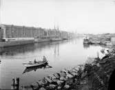 En bild från inre Gävle Hamn. Roddbåten August med rorsman är i tjänst. Ångbåten Freja väntar på avgång

