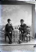 Gruppbild, två män sittande i helfigur vid ett litet trekantigt bord. På bordet två pilsnerflaskor och männen håller i varsitt glas. Männen bär hatt. Ateljébild. Beställare: Smed  A.V. Lindman.