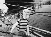 Liten pojke med en timmerstör, vid sjökanten.