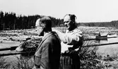 Två män vid älv kanten, tittandes på timmerstockar.